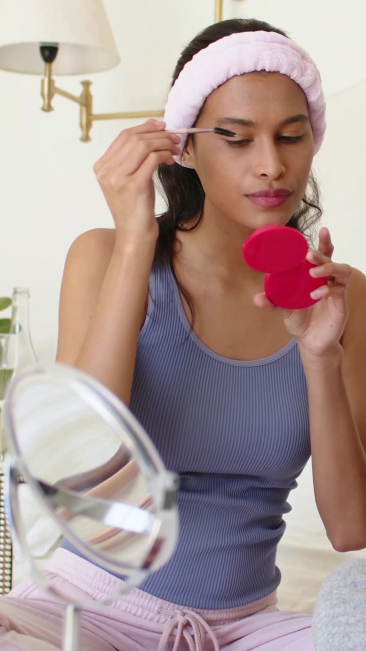 Vertical video: Leaning woman pink headband at home reaching mirror grooming brows with red compact
