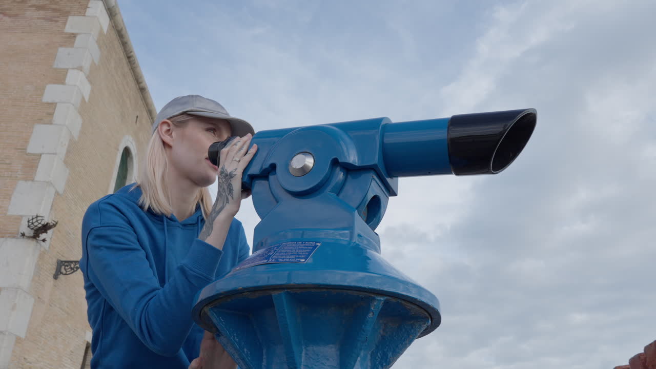 mujer rubia mira a través del telescopio hacia edificios históricos y el cielo, estudiante turista haciendo turismo