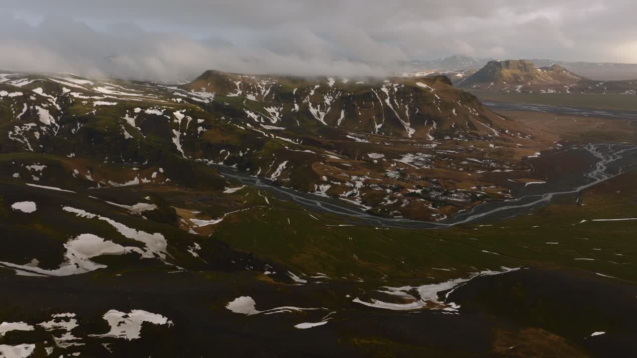 vista panorámica aérea de un río que fluye a través de montañas cubiertas de nieve derretida, en un día nublado y brumoso, en islandia