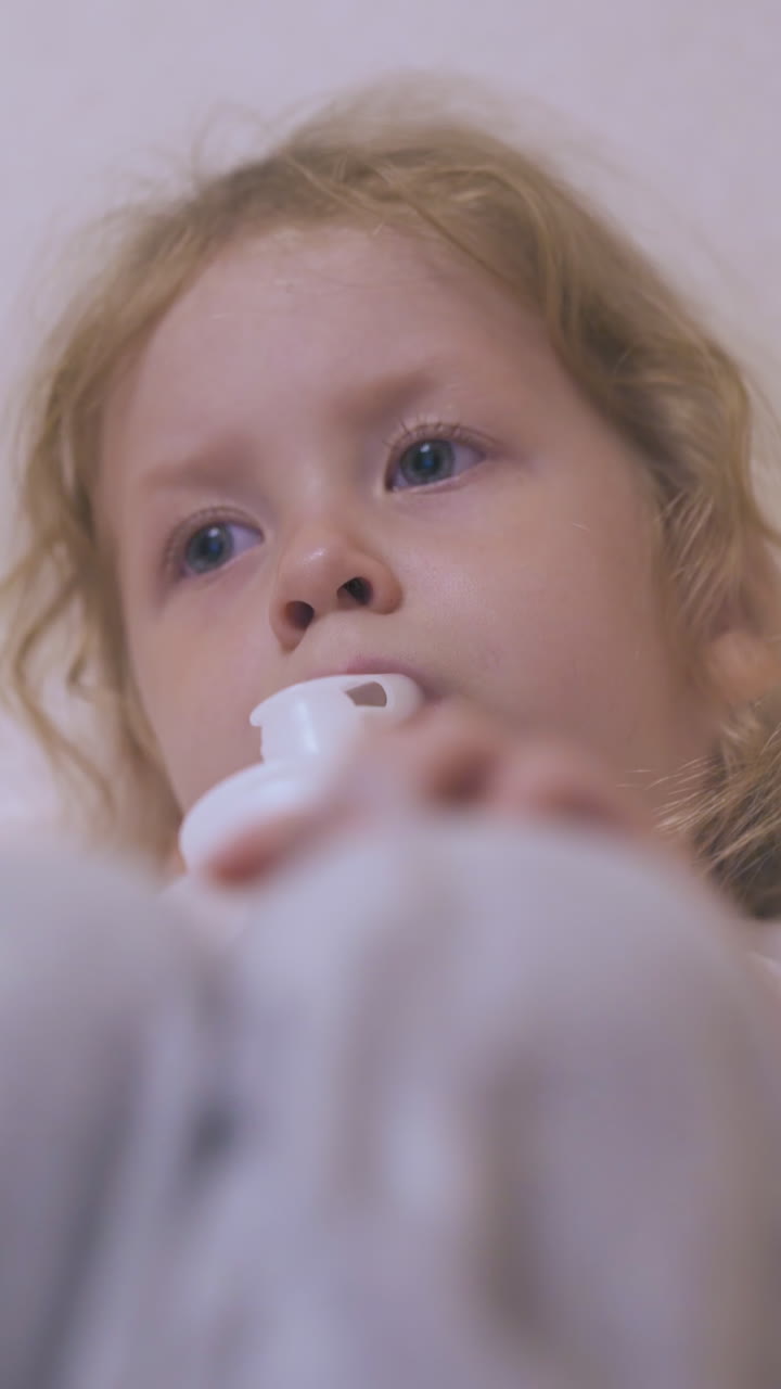 adorable blue eyed girl has procedure with modern inhaler lying in soft bed in light room at home focus on knees