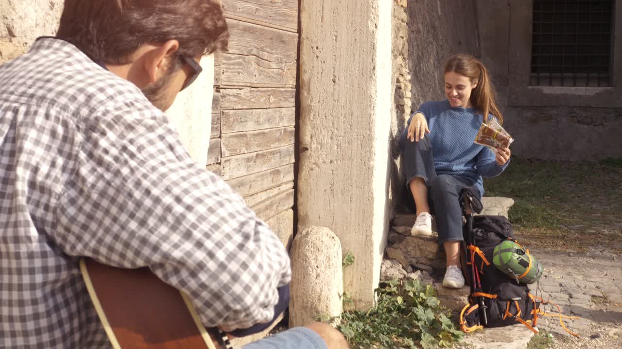 pareja feliz de mochileros turistas tocando la guitarra y cantando sentados cerca de la puerta de madera de un edificio medieval en el parco degli acquedotti ruinas del parque en roma al amanecer con guía de mapa cámara constante en cámara lenta