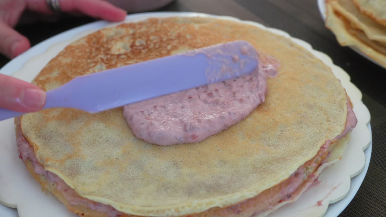 Close-up shot of hands skillfully spreading a luscious berry filling onto a golden crepe. The soft texture and gentle lighting create a cozy kitchen atmosphere, perfect for cooking enthusiasts