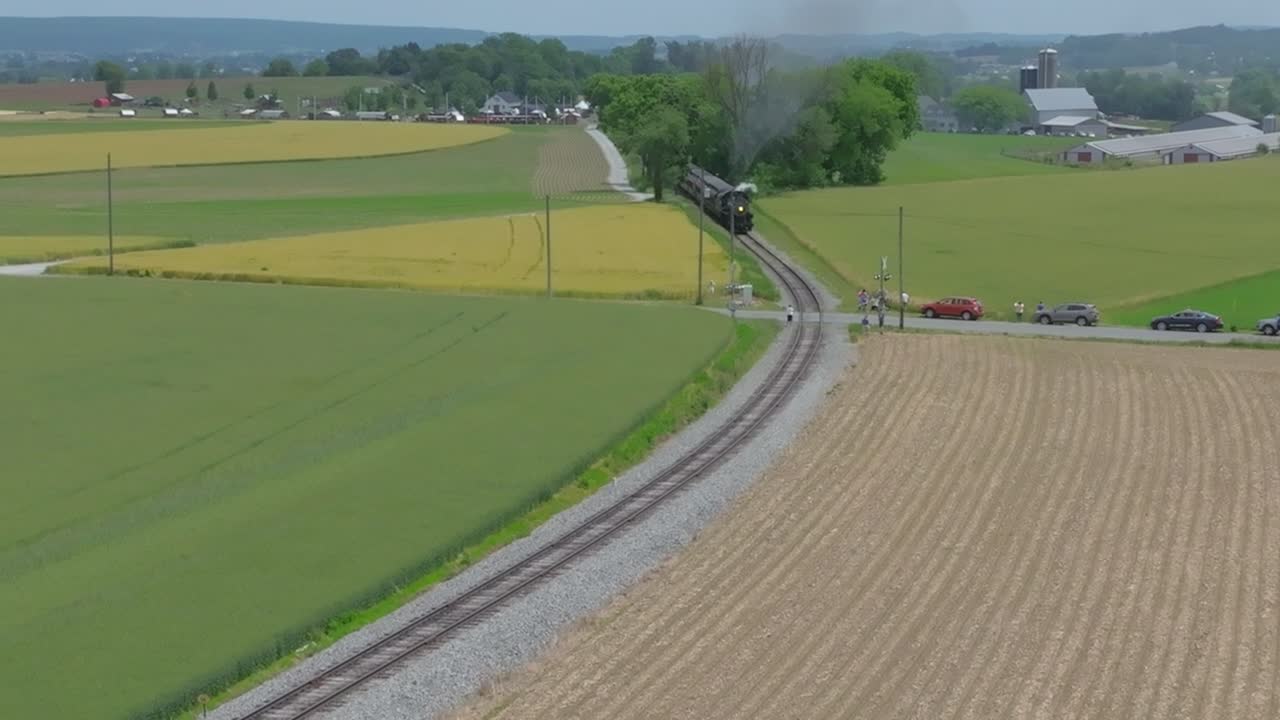 A train smoothly travels along the tracks surrounded by expansive fields and trees. Cars pass on nearby roads, showcasing the tranquil rural setting and distant buildings under a clear sky