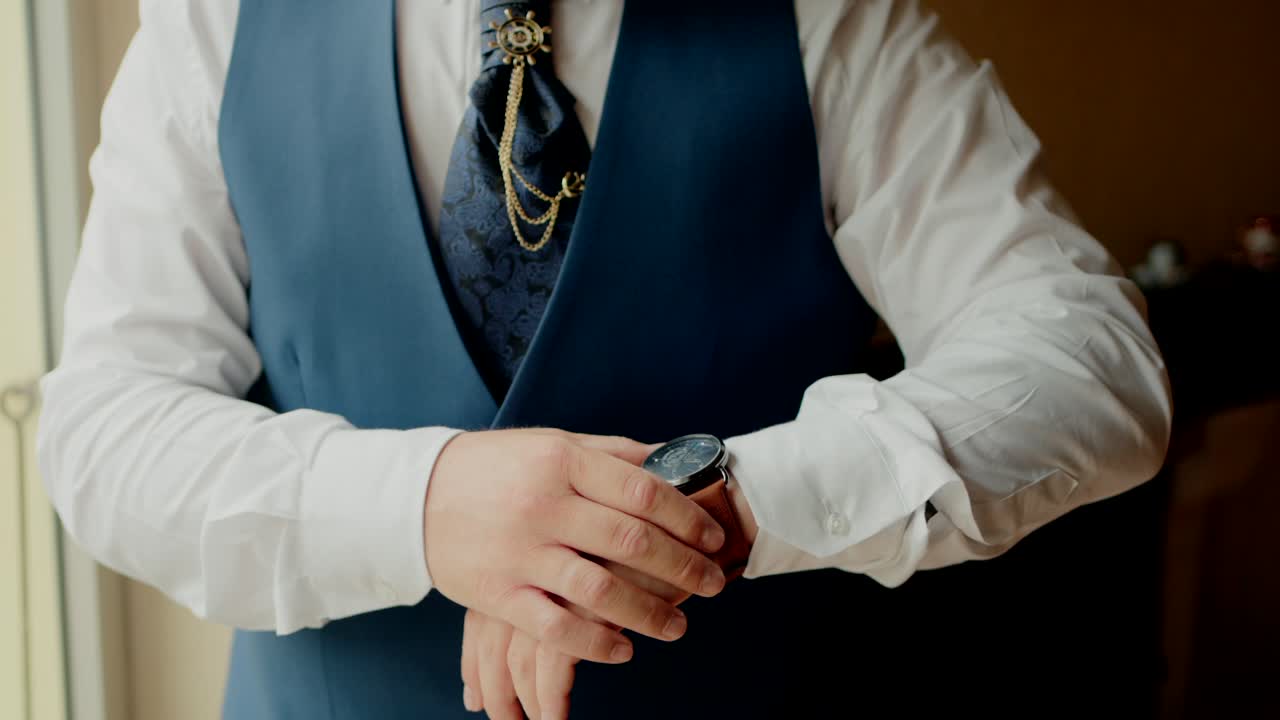 groom adjusting his watch while wearing a blue vest, gold chain, and tie