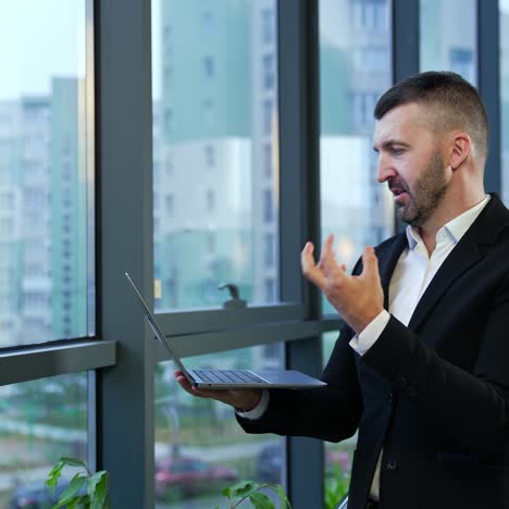 male entrepreneur standing at the window and holding laptop in front of him. business talk on the computer. man talking and gesturing in front of notebook