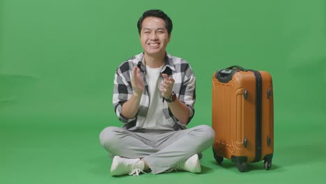 full body of asian male traveler with luggage smiling and clapping his hands while sitting in the green screen background studio