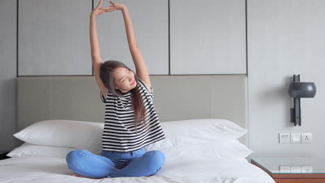 pretty young smiled asian woman stretching her hands up while sitting on a bed with crossed legs in the morning