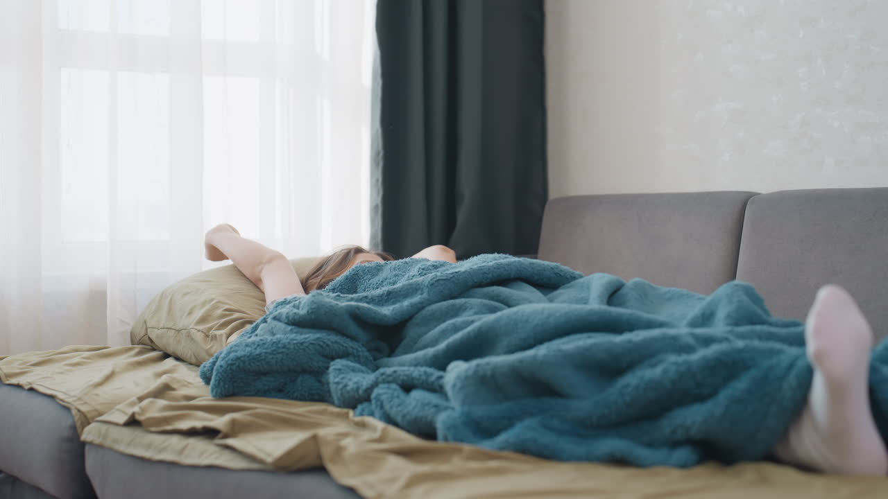 Woman Freelancer Reaching While Lounging Under Teal Blanket On Sofa, Arms Extended Toward Morning Light In Minimalist Living Room Relaxed SlowStart Scene Blending Rest And Remote Work Balance
