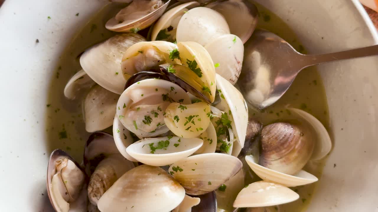 Close-up of clam soup with garlic and parsley in a bowl, captured in natural lighting at a beachside restaurant