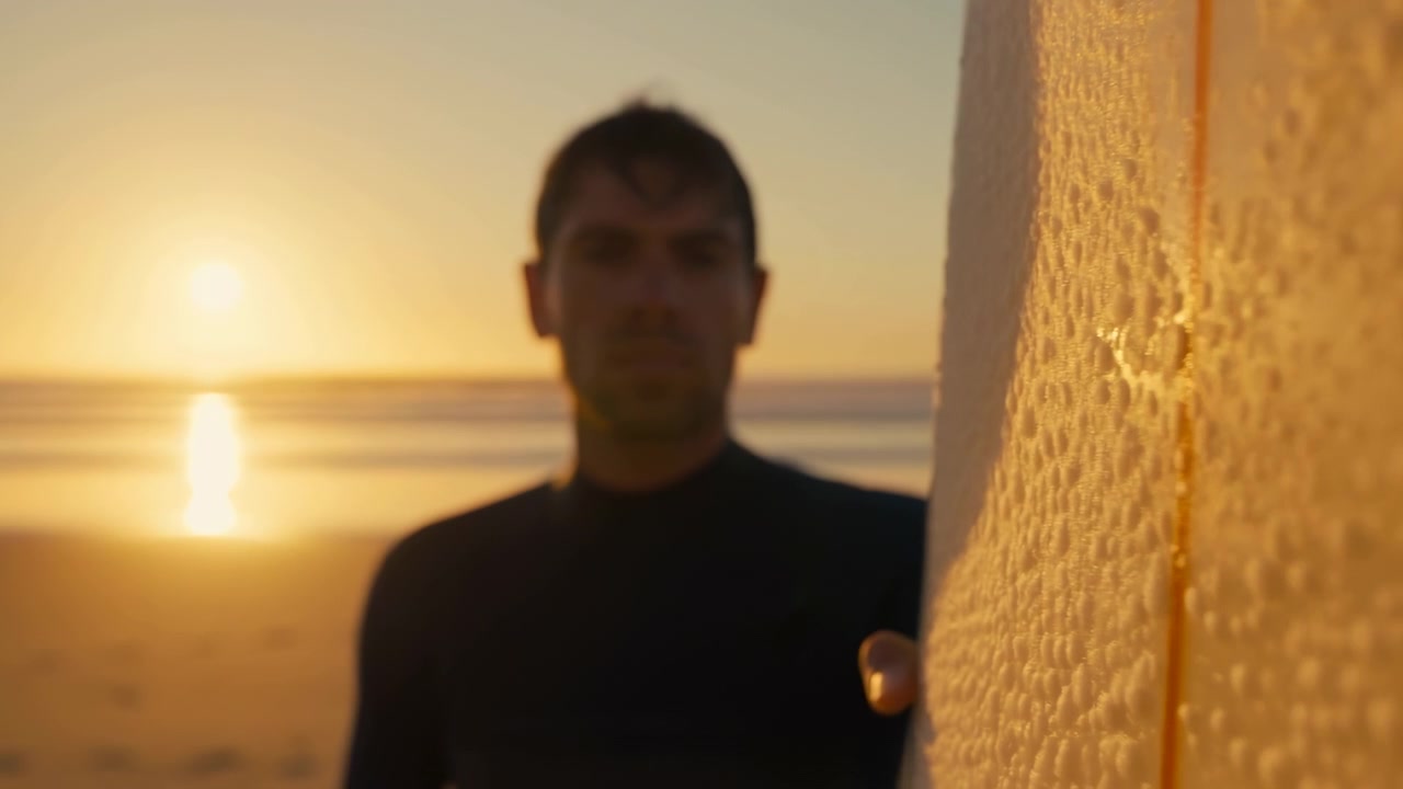 Una persona borrosa y silueteada está de pie junto a una superficie reflectante texturizada con la cálida luz del atardecer brillando sobre agua tranquila en el fondo, creando un ambiente suave.