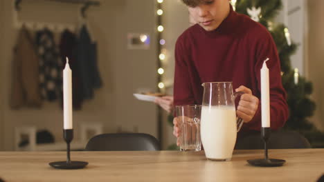 un niño rubio y su hermana rubia colocan un vaso vacío, una jarra de leche y un plato lleno de galletas en una mesa vacía con dos velas, luego salen de la habitación