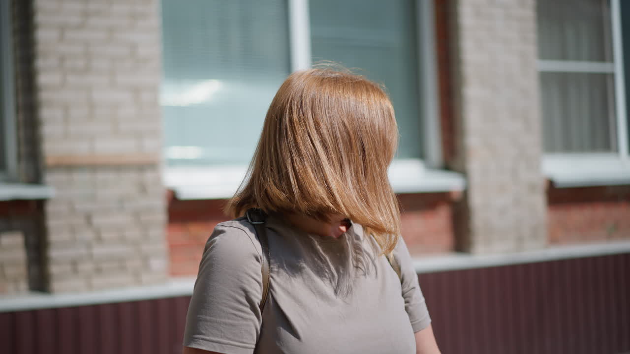 Medium view of college student standing under bright sunlight near school building, looking around and checking time with fitness bracelet, showing sadness and exhaustion on warm summer day
