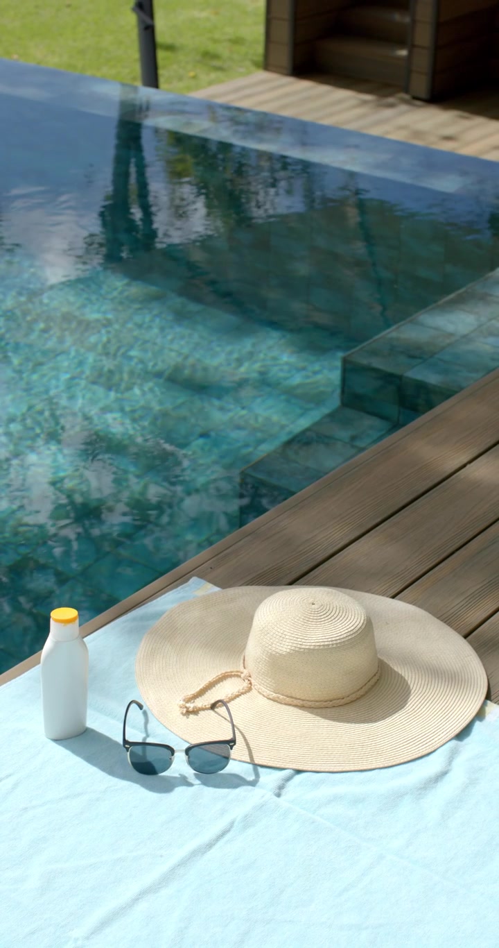 Vertikales Video: Leichte Brise kräuselt den Pool, während Hut, Sonnenbrille und Sonnencreme auf einem Handtuch liegen.