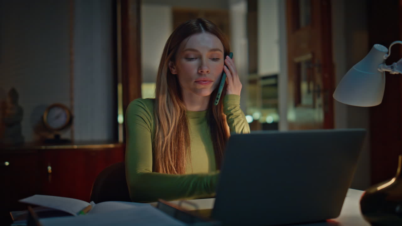 Late worker talking cellphone sitting at remote workplace with laptop close up.