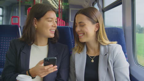 two businesswomen with takeaway coffees commuting to work on train looking at mobile phone together