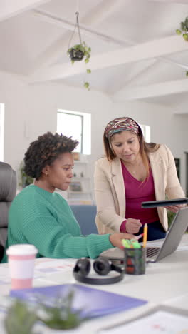 vertical video of diverse business female colleagues discussing using laptop in office, slow motion