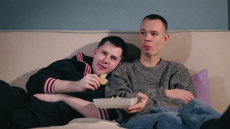 two men watching movies and eating chips on a couch