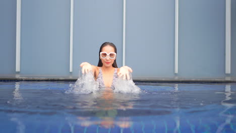 asian woman inside swimming pool wearing an white sunglasses splashing water with both hands