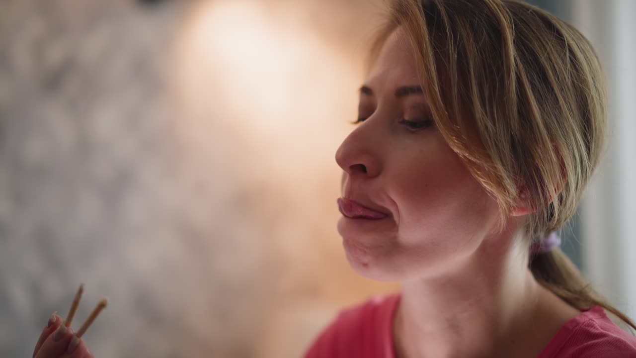 Close up of lady in pink top eating happily and licking chopstick after savoring meal while seated in cozy indoor setting with soft blurred background