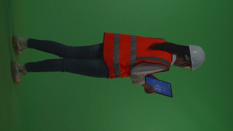 back view of full body asian female engineer with safety helmet looking at the wind turbine blueprint on a tablet and looking around while standing in the green screen background studio, during sunset or sunrise time
