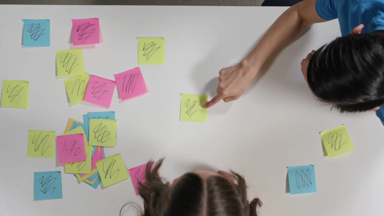 Noticing loose pile, two children wearing yellow and blue sorting neon sticky notes at white table