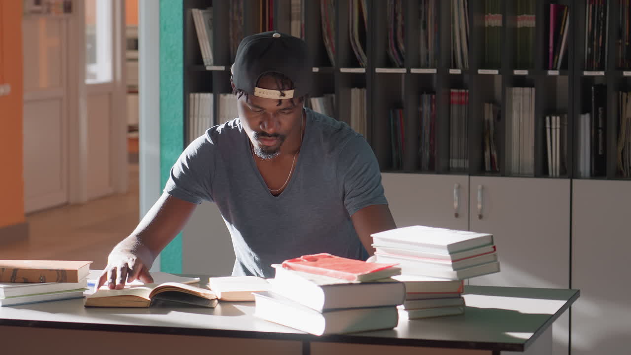 high school student in casual cap studies surrounded by stacks of books in bright library, tapping finger thoughtfully on open book under sunlight, showing concentration and academic dedication