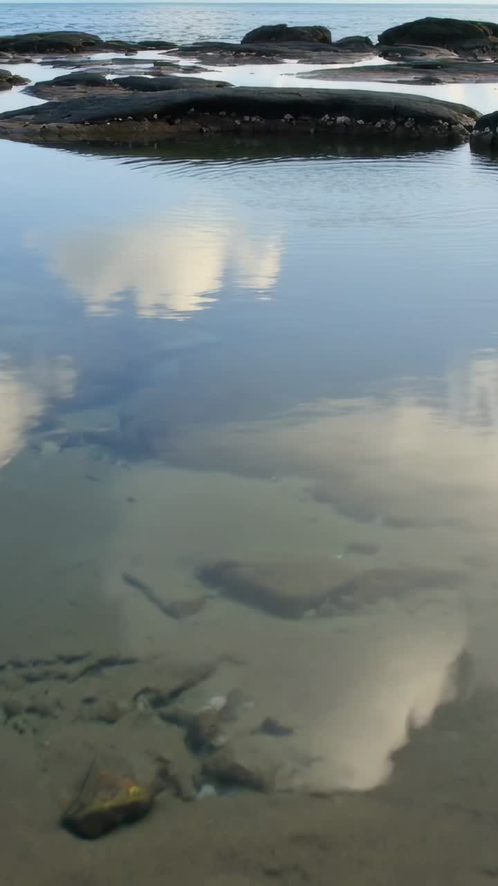 Vertical video: Passing cloud nudging shallow tide pool, showing submerged stones near rock ledge