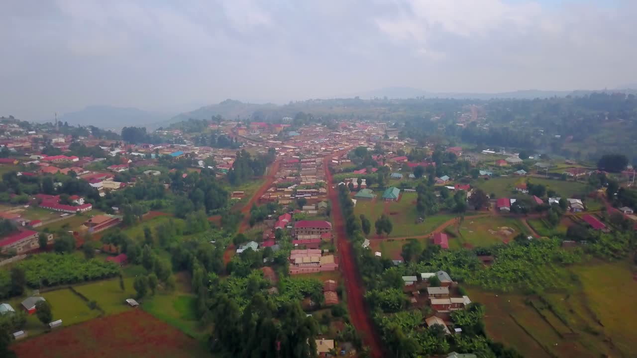 Drone footage over Kapchorwa town in Uganda shows a central red dirt road lined with houses and trees, surrounded by fields, hills, and lush vegetation under a cloudy sky in the elevated highlands