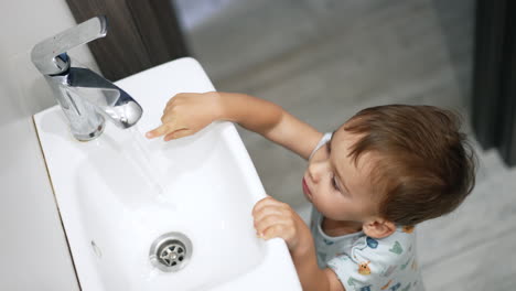 cute toddler stands holding by the sink. adorable child touches the water flowing from the tap. top view.