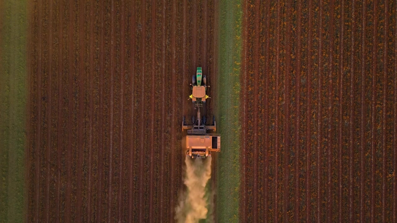Ripresa aerea dall'alto con tracciamento di un trattore che ara i solchi in un campo agricolo, producendo una scia di polvere mentre si muove.
