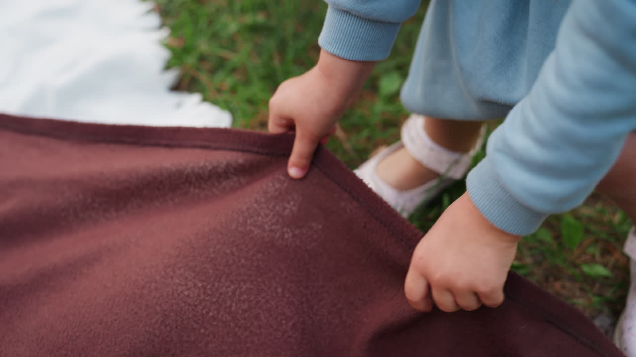 평화로운 야외 피크닉 준비 순간에 초점을 맞추고 세심하게 작업하는 작은 손을 보여주는, 녹색 잔디 위의 흰색 매트 위에 갈색 천을 조심스럽게 놓으면서 피크닉 천을 정리하는 유아의 항공 뷰