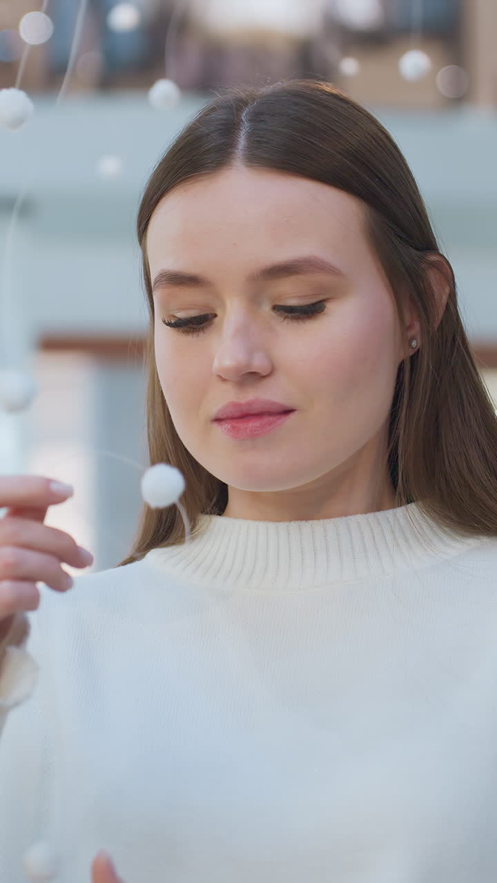 Young lady in elegant white sweater stands in festively decorated shopping mall, gently holding delicate hanging ornament, surrounded by soft lights, she admires the festive ambiance