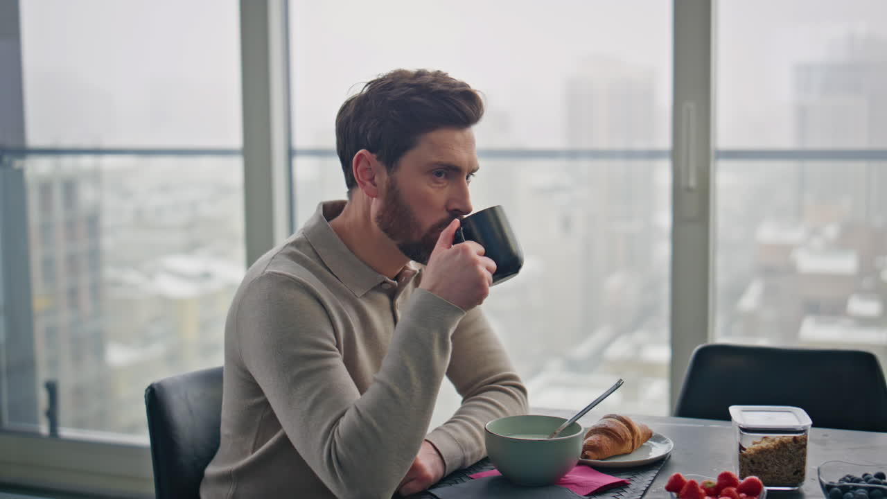차분한 남자가 커피 컵을 마시며 럭셔리 주방 테이블에 앉아있는 클로즈업.