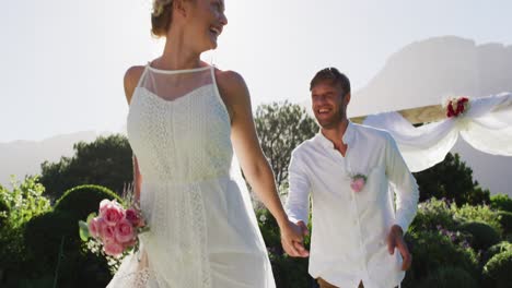 Portrait-of-happy-caucasian-newly-wed-couple-running-from-altar-outdoors