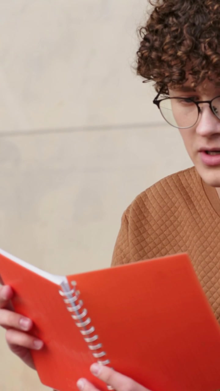 Video verticale: Un uomo in maglione solleva un quaderno arancione, studia e poi lo chiude su una panchina del campus.