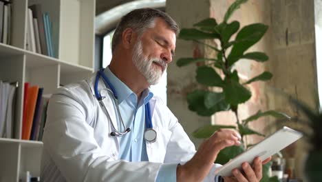 senior doctor thinking about patient's diagnosis and typing on a tablet computer. his office is bright and modern.