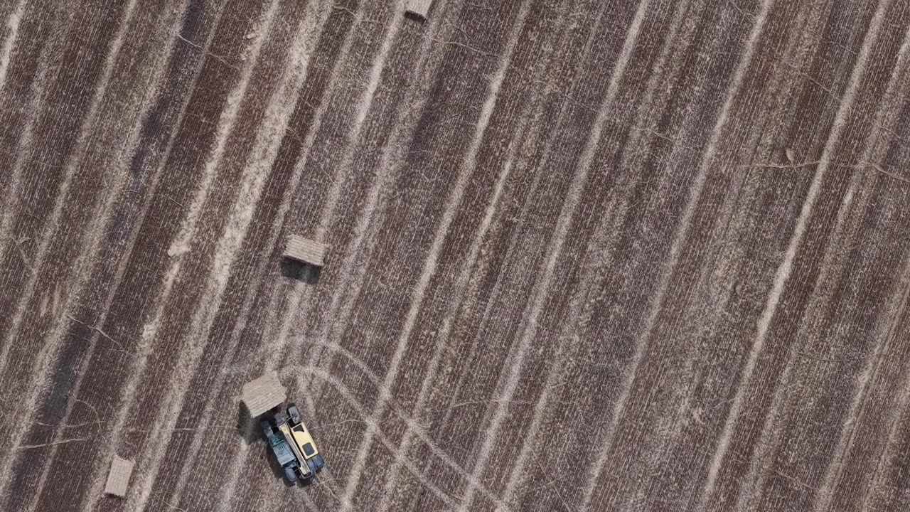 Una veduta aerea riprende un trattore giallo che imballa attivamente il fieno in un vasto campo agricolo, mettendo in mostra operazioni agricole moderne ed efficienza