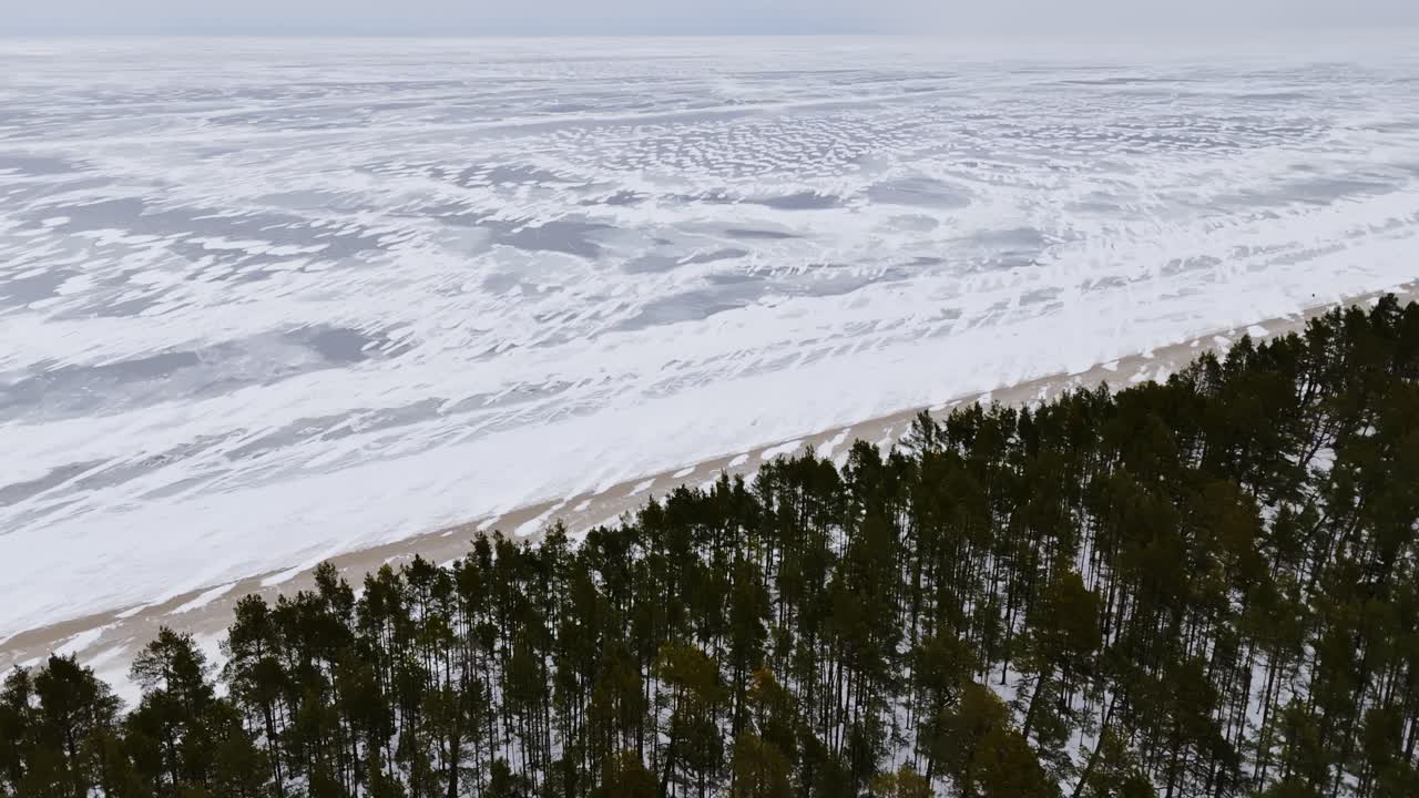Вид сверху с дрона на замерзшее море с узорами снега, наметанными ветром, и узким песчаным пляжем рядом с темным сосновым лесом. Чистый зимний фон побережья с большим пространством для текста