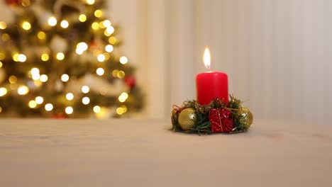 single red festive christmas candle decoration burning and sitting on table with shallow focus decorated christmas tree with lights on in the background