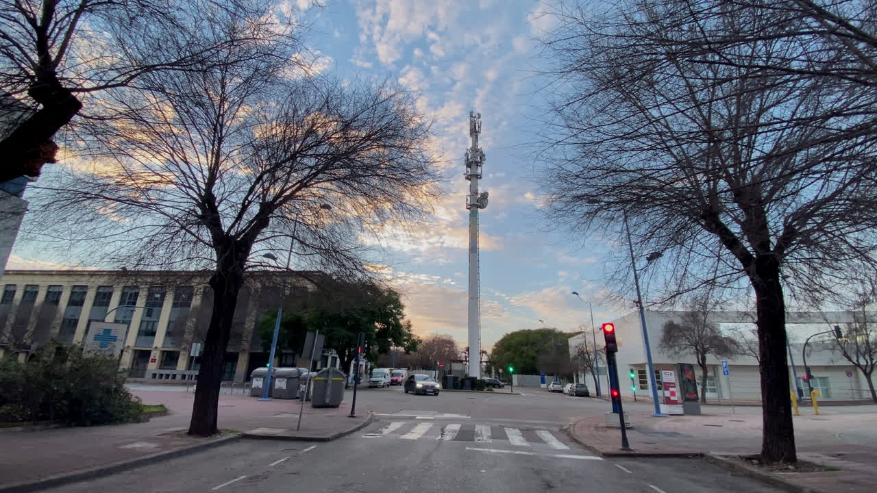 Scène de rue urbaine avec une antenne-relais mobile, des arbres nus et des bâtiments sur fond de ciel de soirée nuageux à Séville.