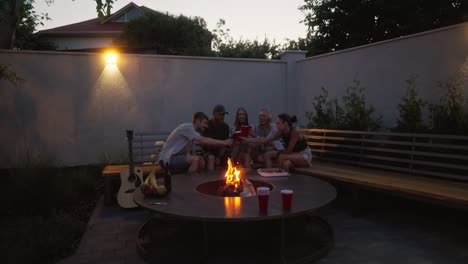 Side-view-of-a-happy-group-of-cheerful-friends-clinking-their-glasses-while-sitting-on-a-bench-near-a-fire-talking-and-having-fun-during-their-holiday-in-a-country-house