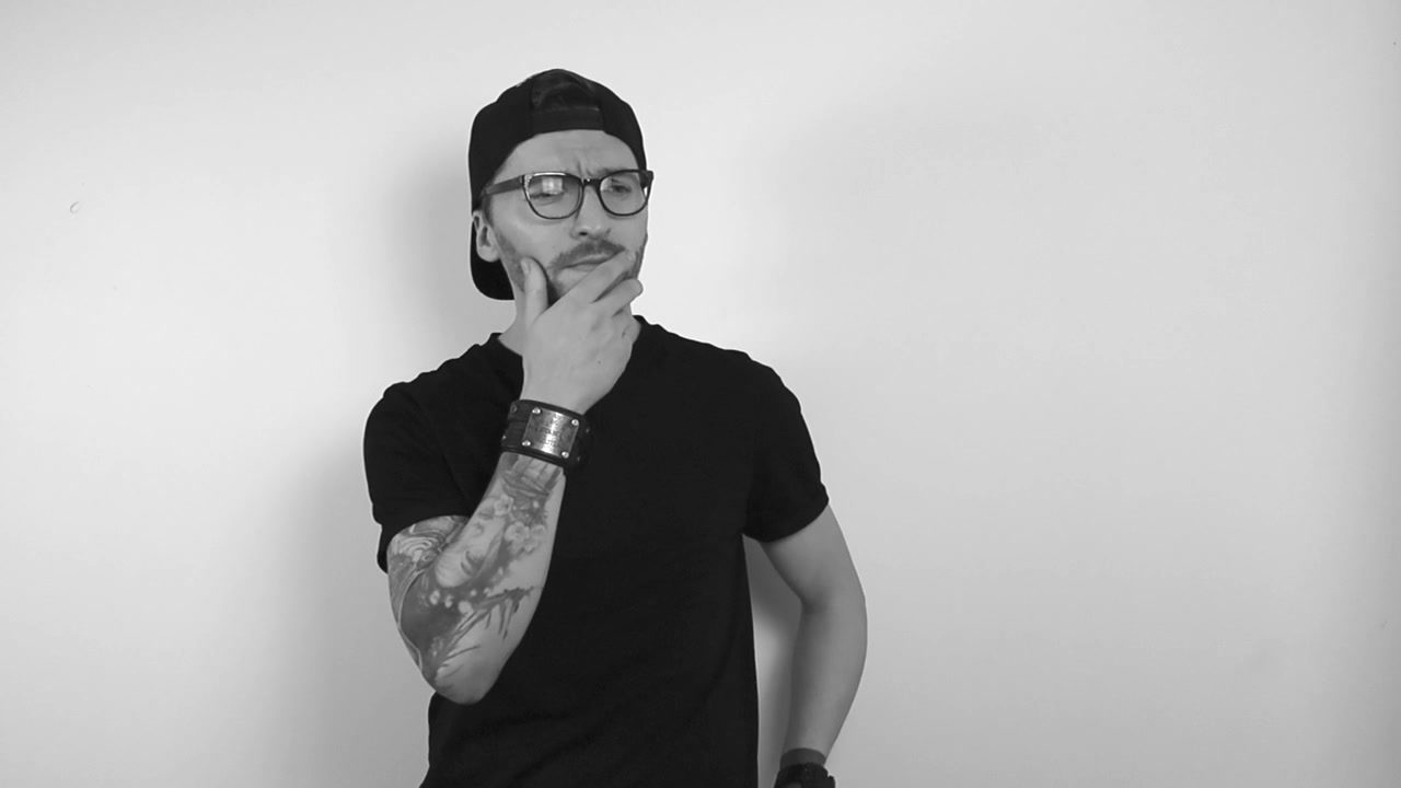 Filmato in bianco e nero di un uomo con un tatuaggio a manica intera che non riesce a vedere qualcosa e indossa occhiali con montatura nera. Inizia a strizzare gli occhi e ad avvicinarsi per vedere meglio, vede e annuisce in seguito