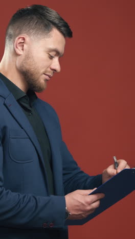 half-length video of handsome young smiling businessman wearing black shirt and dark blue suit holding the folder taking notes. dark red background. vertical video