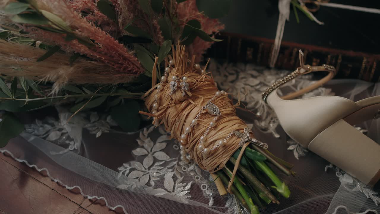 bridal bouquet wrapped in straw, pearls, and lace rests on a veil beside elegant heels