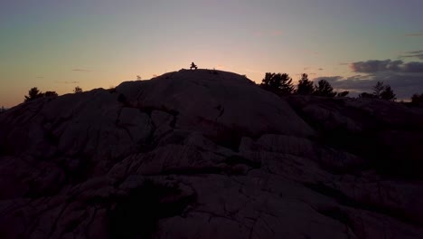fly over inukshuk on rocky pine tree island at sunset, drone aerial wide dolly in