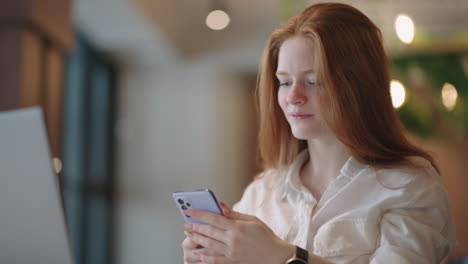 Red-haired-Woman-working-from-home-using-laptop-computer-while-reading-text-message-on-mobile-phone.-Woman-using-a-phone.-Serious-charming-woman-using-smartphone-while-working-with-laptop-at-home