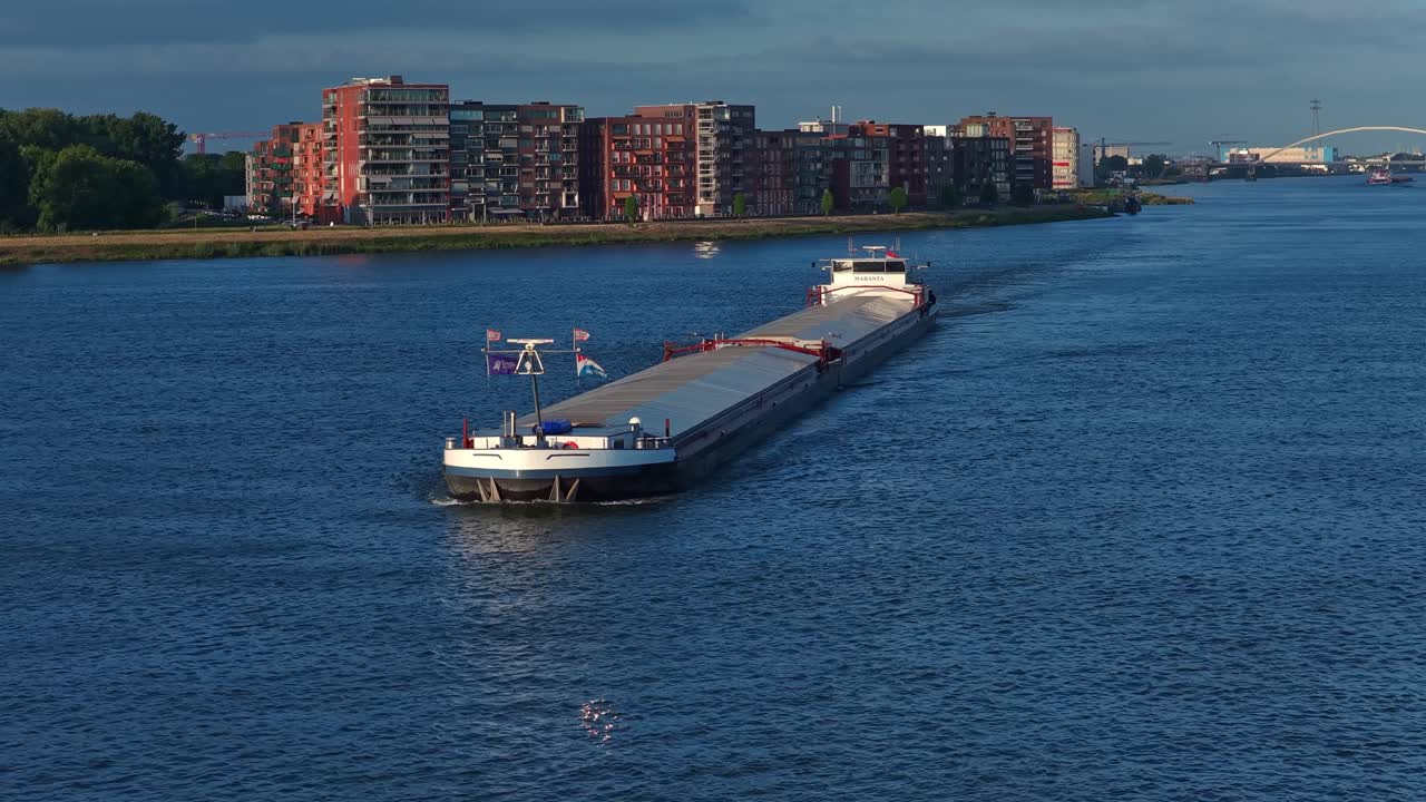 The drone flies forward toward a cargo ship on a wide canal in Dordrecht, with residential buildings, quays, and clear blue sky in the background