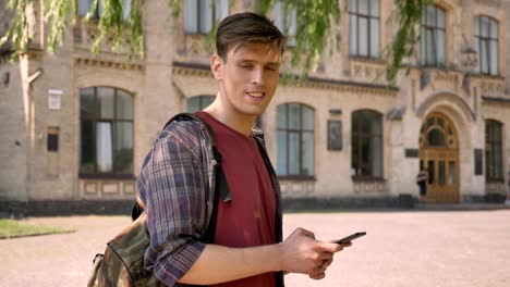 joven apuesto está escribiendo en su teléfono inteligente en el parque durante el día en verano, mirando a la cámara, concepto de comunicación