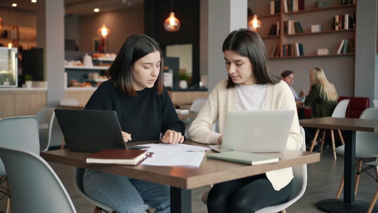 Kvinne peker mot venstre og viser linjer for en kvinne til høyre som leser papirer, skriver på bærbare datamaskiner og studerer på en kafé.