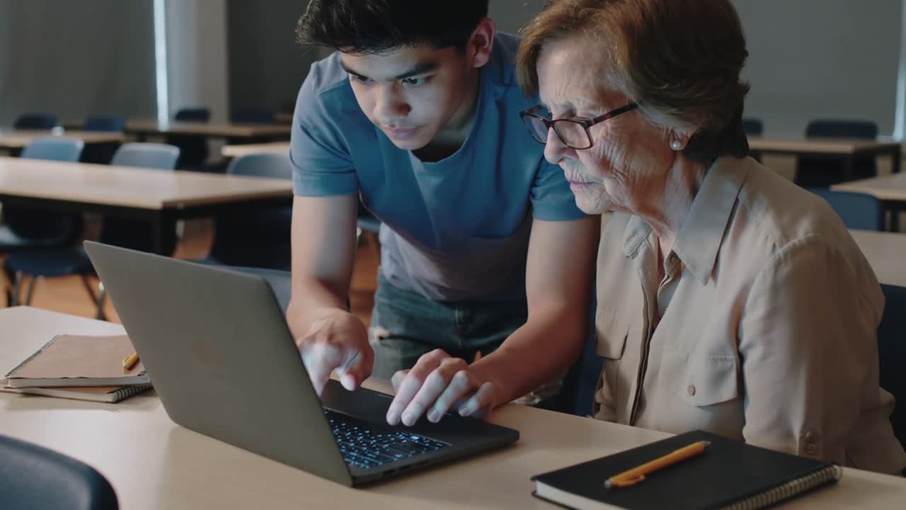 Seeing laptop, teen male in blue tee teaching senior woman with glasses, typing pointing in class